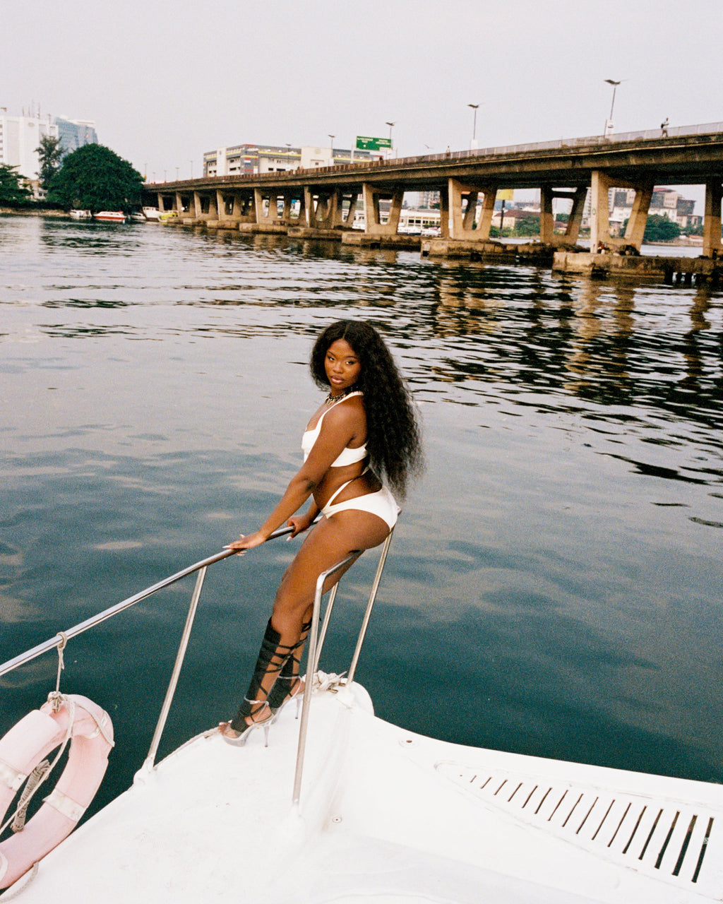 Woman in a white Herve Leger bandage one-piece swimsuit sitting on a boat with a cityscape in the background